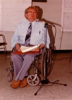 Henry Enns sit in a wheelchair front opf a micro and with papers on his knees.